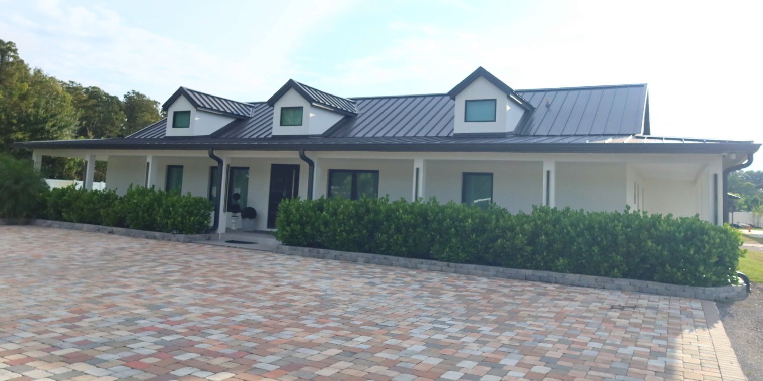 Dark standing-seam metal roof with three dormers on a white single-story home with a wraparound porch.